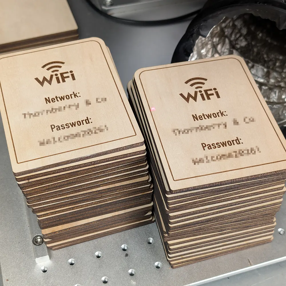 A production run of laser-engraved birch Wi-Fi signs stacked on the engraving bed, each engraved with a network name and password.